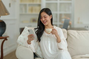 Asian woman in white pajamas sitting on the sofa looking at mobile phone online, holding a cup of hot coffee. Smiling happily in her living room on a relaxing morning. Holiday concept, lifestyle.