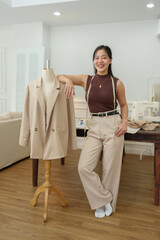 Asian tailor woman works on clothes in a tailor shop. Attractive beautiful young designer smiles after successfully making a suit on a mannequin in the office with a laptop design concept.