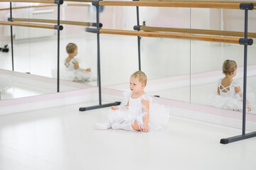 Portrait of a cute baby ballerina sitting near the ballet barre putting on shoes