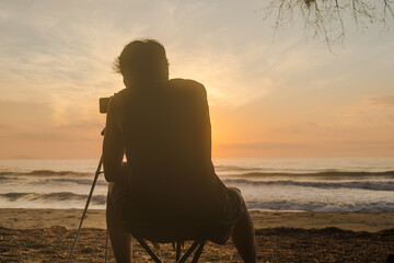 silhouette phone on tripod and man sit on beachchair during record vdo and photo beach with sunrise...