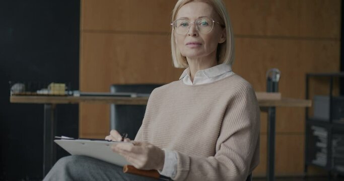 Portrait of experienced female psychologist writing then looking at camera sitting in armchair in office. Therapy and professionnal occupation concept.