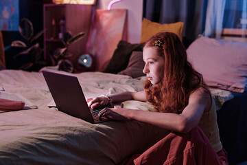 Medium shot of red-haired teen girl sitting on floor in bedroom late in evening surfing Internet on...