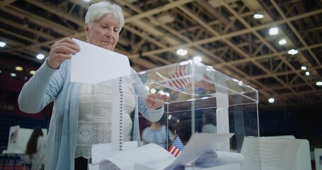 Close up shot of polling box standing on table in polling station. Elderly female voter puts bulletin into box, looks at camera. Political races of US presidential candidates. National Election Day.