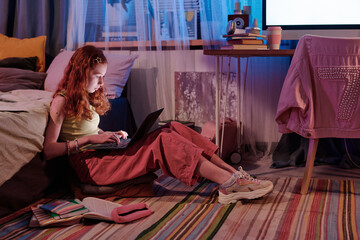 Side view of modern teen girl spending evening in her bedroom, sitting on floor and typing on...