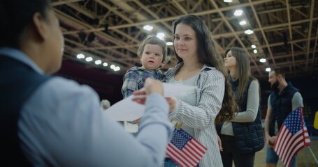 Woman with baby on hands talks with polling officer and takes bulletin. Female American citizen comes to vote in polling station. Political races of US presidential candidates. National Election Day. - obrazy, fototapety, plakaty