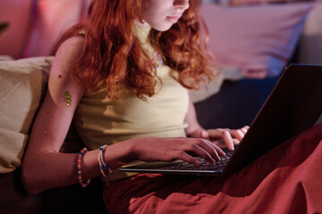 Medium section crop shot of red-haired teen girl sitting in bedroom typing on laptop late in evening