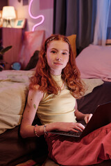Vertical medium shot of red-haired girl wearing casual clothes sitting in bedroom looking at camera...