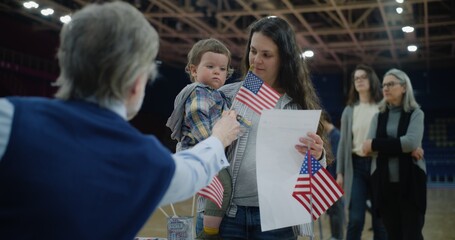 Woman with baby on hands talks with polling officer and takes bulletin. Female American citizen comes to vote in polling station. Political races of US presidential candidates. National Election Day. - obrazy, fototapety, plakaty