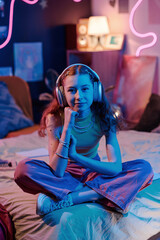 Vertical shot of stylish teen girl wearing headphones sitting on bed in her bedroom decorated with...