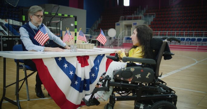 Woman with spinal muscular atrophy in electric wheelchair consults with worker in polling station. Political races of US presidential candidates. National Election Day in United States. Dolly shot.