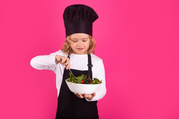 Funny child cook hold plate with vegetables. Kid in cooker uniform and chef hat preparing food on studio color background. Cooking, culinary and kids food concept.