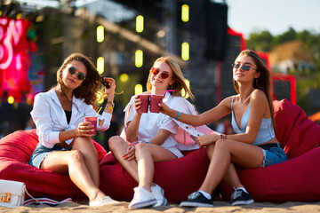 Girls in sunglasses sit on red bean bag chairs, cheer with drinks at beach music festival. Trio...
