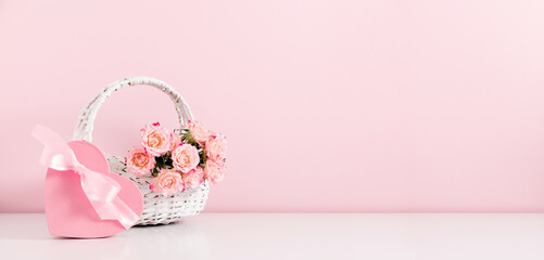 Beautifull bouquet of pink rose flowers in a wicker basket and a gift box with satin bow on pastel pink table background. Birthday, Wedding, Mother's Day, Valentine's day, Women's Day. Front view