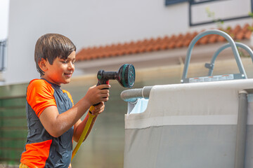 Cheerful boy with wet hair filling up a pool using a garden hose, on a sunny day.