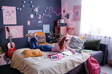 Wide shot of red-haired teen girl wearing headphones lying on bed in her bedroom making notes...