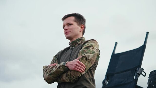 From the side a confident man with a short haircut in an army camouflage uniform stands confidently against the sky and looks to the side with his arms folded on his chest outside the city