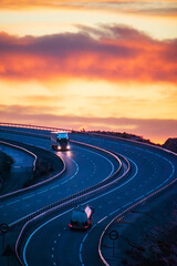 Two trucks driving in opposite directions on a highway with curves and a dramatic sky at dawn, a...
