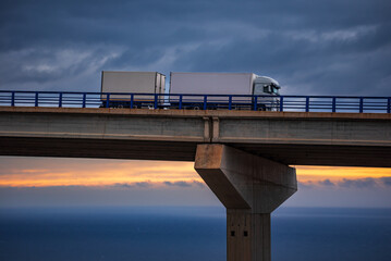 Three-axle truck with trailer driving along a viaduct, with the sea on the horizon and a dawn sky.
