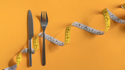 Fork and knife wrapped with measuring tape on yellow background. Weight loss and healthy concept