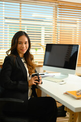 Portrait of successful businesswoman with cup of coffee sitting at desk and smiling at camera
