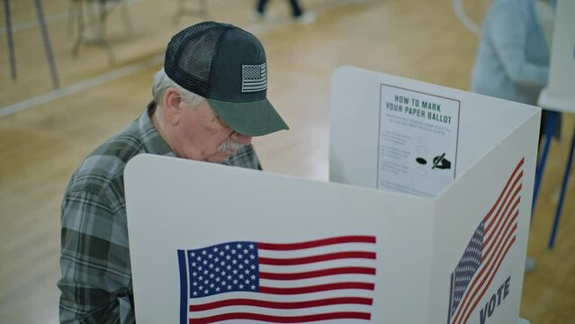 Elderly male voter with bulletin in hands comes to voting booth. American citizens come to vote in polling station. Political races of US presidential candidates. National Election Day. Dolly shot.