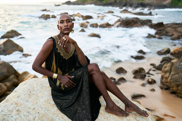 Non-binary ethnic fashion model in dress, brass jewelry sits on rocks by ocean. Trans sexual black person with rings, nose-ring, bracelets, earrings in posh clothes poses in tropical seaside location.