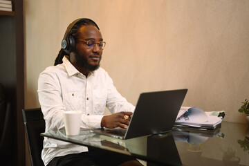 Smiling African millennial guy in headphone working remotely on laptop at home