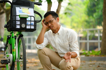 Upset male worker sitting near broken bicycle in city park during go to work. Transport concept