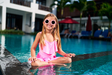 Smiling girl in pink swimsuit enjoys summer by pool. Happy child with sunglasses sits at water...