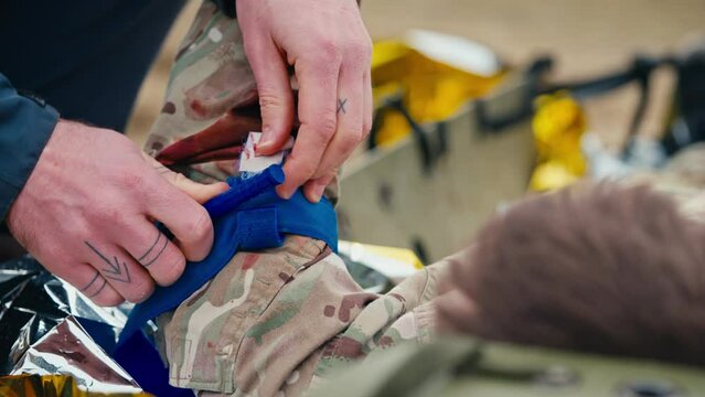 Close up a confident male medic in a dark blue uniform puts a bandage on the wounded hand of a soldier to avoid blood loss during combat operations outside the city