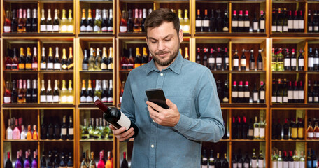 Digital marketing in retail. Man consumer stands in liquor store holds wine bottle in hand and using his smartphone.