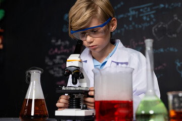 Blonde hair schoolboy in laboratory wear lab coat stand and learn science of chemistry technology in STEM class. The student looking object in microscope . On the table put many flask. Erudition.