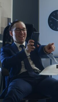 Medium vertical of toxic career man in business suit, tie and glasses sitting in chair at desk, using smartphone, tripping up colleague walking by with documents, pointing finger, laughing viciously
