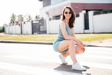 Young beautiful smiling female in trendy summer blue cycling shorts and tank top clothes. Carefree woman posing in street. Positive model having fun. Cheerful and happy. Sits on asphalt. In sunglasses