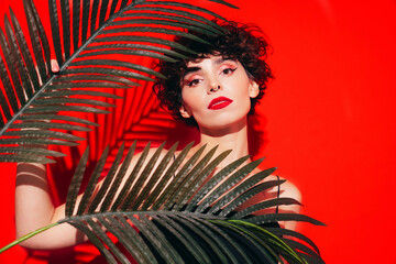 Fashion beauty portrait of young confident brunette woman with evening stylish makeup and perfect clean skin. Sexy model with curly hair posing in studio. With red bright lips. Tropical palm leaf