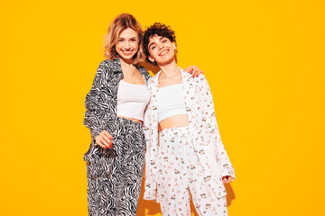 Two young beautiful smiling brunette hipster female in trendy summer costume clothes. Sexy women posing near yellow wall in studio. Positive models having fun. Cheerful and happy