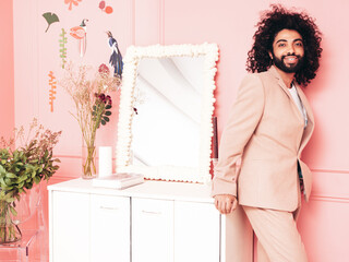 Handsome smiling hipster  model. Unshaven Arabian man dressed in elegant beige suit . Fashion male with long curly hairstyle posing near pink wall in pure interior with flowers in studio