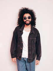 Handsome smiling hipster  model. Sexy unshaven Arabian man dressed in summer stylish black jacket and blue jeans. Fashion male with long curly hairstyle posing near grey wall in studio