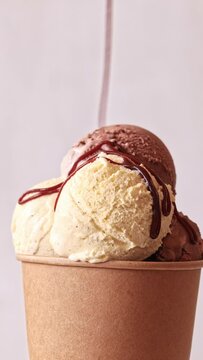 Chocolate sauce flowing on Chocolate and vanilla ice cream scoops in paper take away cup rotating over light background. Sweet dessert, rotation closeup. 4K