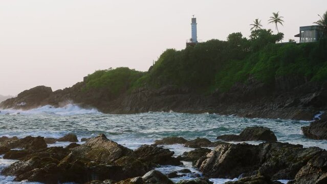 Lighthouse stands on green hill above ocean waves crashing against rocky shore at sunset. Tranquil dynamic nature, meditation, scenic backgrounds. Sunlight glistens water, serene dusk coast landscape.