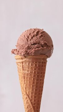 Close up of chocolate flavor ice cream ball in waffle cone rotating over light grey color background. Summer dessert