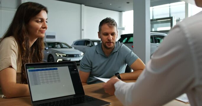 A man signs a car purchase agreement at a car dealership. The manager is sitting at his desk in front of an open laptop