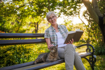 Happy senior woman enjoys reading book and spending time with her cat while sitting on a bench in her garden.	