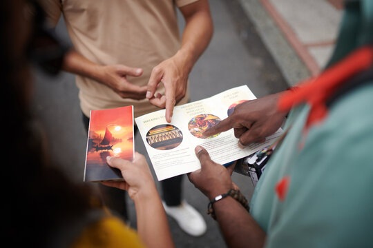 Hands of young male traveler and guide pointing at brochure template during excursion while discussing famous sights of the city