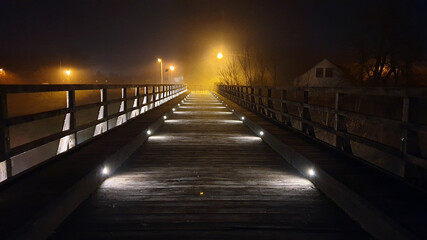 Fototapeta na wymiar Dilapidated old vintage pedestrian wooden bridge with wooden handrails renovated and updated with decorative modern LED lights at night engulfed with thick fog and bright city lights in background