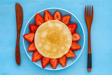 Pancakes and strawberries laid out in the form of flower on blue ceramic plate over wooden...