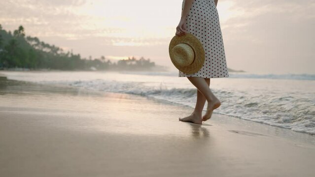Female feet going barefoot in sea surf water edge at sunset. Splashing foamy water. Young woman's legs walk slowly along ocean shore at summer evening. Lady in dress enjoying vacation at the seaside.