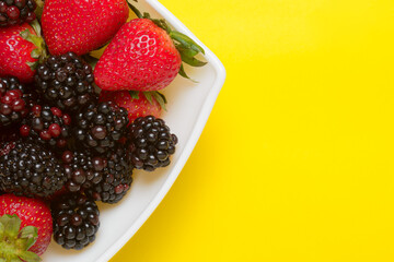 Blackberries and strawberries in a bowl on a yellow background. Seasonal fruits. Summer background.