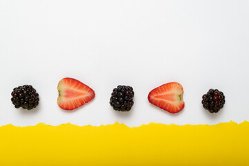 Blackberries and cut strawberries on yellow background with torn white paper border. Summer...