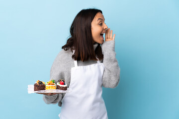 Pastry chef holding a big cake over isolated blue background shouting with mouth wide open to the...